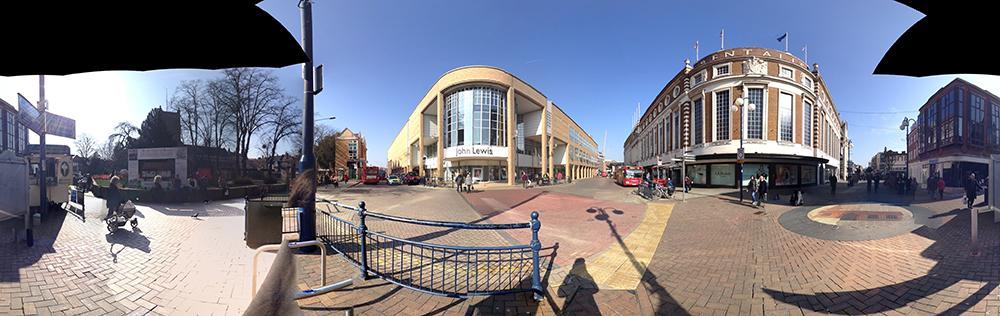 A panoramic image of a high street in England. There is a blue sky at the top, two shopping centre buildings in the middle and a brick road at the bottom.